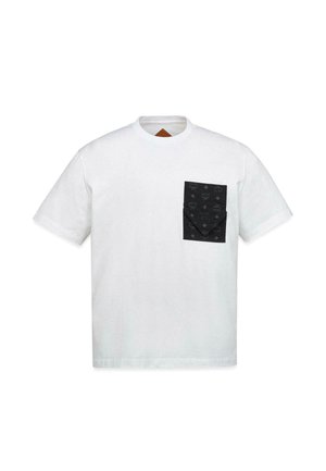 Giovane uomo che indossa una t-shirt bianca con una tasca nera sul petto sopra una camicia a maniche lunghe con fantasia e pantaloni neri larghi, in piedi davanti a uno sfondo semplice.