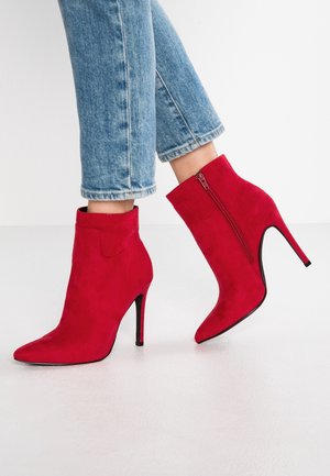 Bottines en suède rouge avec un bout pointu, un talon aiguille haut et un détail de fermeture éclair sur le côté. Portées avec un jean bleu clair retroussé.