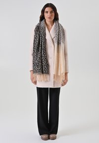 Cappotto bianco in misto lana con ampi rever e due bottoni, abbinato a una sciarpa nera e beige a fantasia con frange e pantaloni neri a gamba larga.