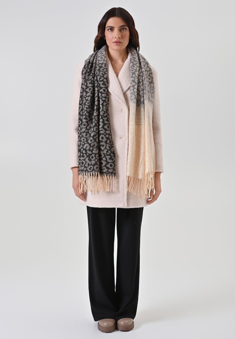 Cappotto bianco in misto lana con ampi rever e due bottoni, abbinato a una sciarpa nera e beige a fantasia con frange e pantaloni neri a gamba larga.