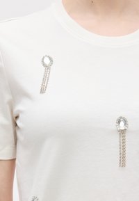 Λευκό πουκάμισο με δύο οβάλ κρυστάλλινες καρφίτσες, η καθεμία με τρεις κρεμαστές σειρές από μικρές στράς, φορεμένες στην περιοχή του στήθους.