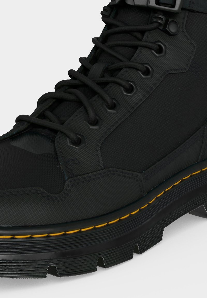 Black rugged boot with thick sole, yellow stitching along the sole, and black laces threaded through metal eyelets.