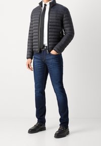 Vadderad svart jacka, vit skjorta, svart slips, blå jeans och svarta loafers med guldaccent. Släta texturer och skräddarsydd passform.