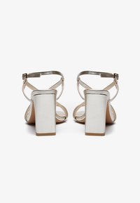 Silver heeled sandals with a block heel, open back, crisscross straps, and a smooth texture. Adjustable ankle straps with metal buckles.