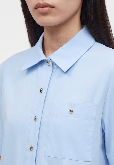 Γυναίκα που φορά ένα γαλάζιο πουκάμισο με γιακά και χρυσά κουμπιά, συμπεριλαμβανομένου ενός στην τσέπη στο στήθος, εμφανίζεται από τον λαιμό έως το μέσο του στήθους.