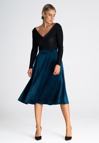 Czarny bodysuit z długim rękawem i dekoltem w kształcie litery V, połączony z turkusową, plisowaną spódnicą do kolan, wykonaną z miękkiego, gładkiego materiału. Całość dopełniają czarne szpilki.