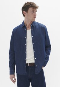 Camisa de mezclilla abotonada en azul oscuro con collar puntiagudo y botones a presión, usada sobre una camiseta de tirantes blanca de canalé. Estilizada con un cinturón marrón.