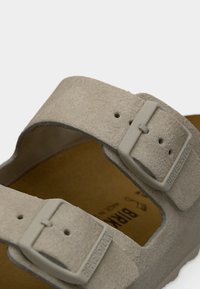 Κοντινή λήψη των λουριών σαμπάνιας Birkenstock από σουέντ με ρυθμιζόμενες αγκράφες πάνω σε καφέ πέλμα.