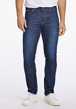 Jeans de mezclilla azul oscuro con corte ajustado, presentan costuras naranjas y una cremallera frontal. Combinados con zapatillas blancas para un look casual.