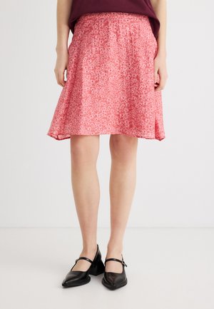 Γυναίκα που φοράει ροζ φλοράλ φούστα μέχρι το γόνατο, σκούρο τοπ και μαύρα πέδιλα Mary Jane, στέκεται μπροστά σε απλό λευκό φόντο.