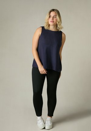 Top senza maniche blu navy realizzato in tessuto morbido con una vestibilità comoda, abbinato a pantaloni neri skinny e sneakers bianche su uno sfondo neutro.