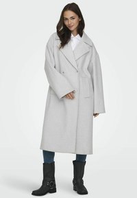 Cappotto in lana grigio, oversize, con ampi revers, bottoni doppi e tasche laterali, abbinato a stivaletti neri e jeans blu.