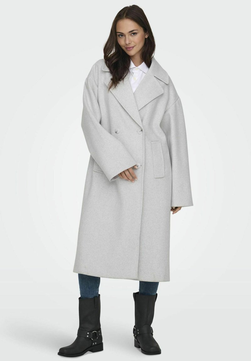 Cappotto in lana grigio, oversize, con ampi revers, bottoni doppi e tasche laterali, abbinato a stivaletti neri e jeans blu.