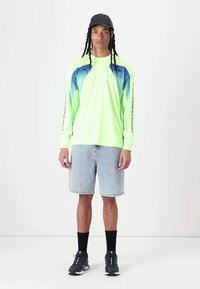 Neon grønn langermet skjorte med blå gradientdetaljer, denimshorts, svarte sokker og svarte joggesko, satt mot en nøytral bakgrunn.