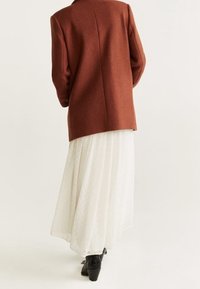 Blazer couleur rouille avec épaules structurées, porté au-dessus d'une jupe blanche plissée fluide à motif à pois et de bottes à talons noires.