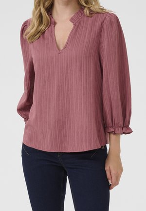 Femme portant un chemisier texturé mauve avec des manches trois-quarts bouffantes et des poignets à volants, associé à un jean bleu foncé, sur un fond uni.