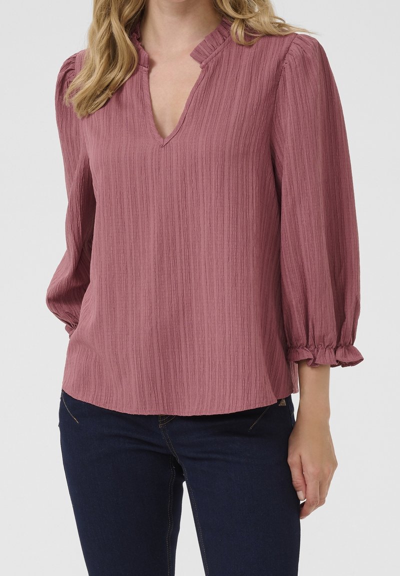 Femme portant un chemisier texturé mauve avec des manches trois-quarts bouffantes et des poignets à volants, associé à un jean bleu foncé, sur un fond uni.