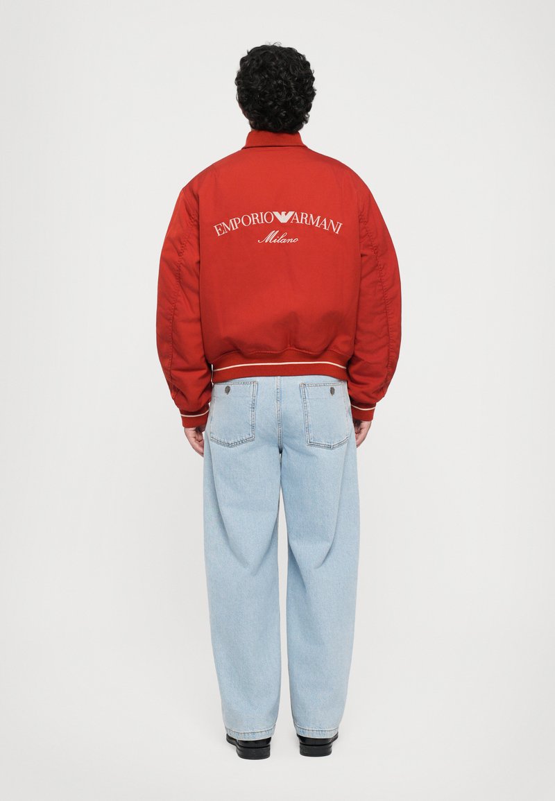 Göndör hajú férfi vörös Emporio Armani Milano dzsekiben és világoskék farmernadrágban, háttal áll egy sima háttér előtt.