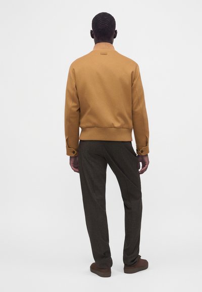 Homme debout de dos portant une veste bomber couleur chameau, un pantalon gris foncé et des chaussures marron sur un fond blanc uni.