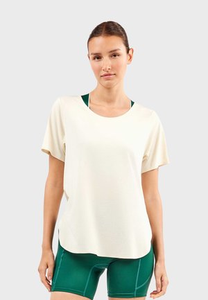 Jonge vrouw met haar in een staart, gekleed in een los off-white topje met korte mouwen en donkergroene aansluitende fietsshorts tegen een effen achtergrond.