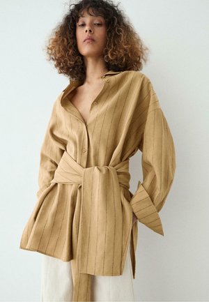 Frau mit lockigem Haar trägt ein übergroßes beiges Nadelstreifenhemd mit breitem, selbstgebundenem Gürtel und weißen Hosen, steht vor einer schlichten hellen Wand.