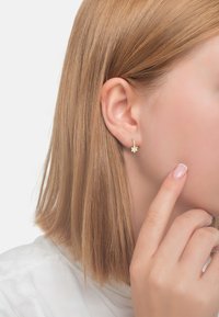 Orecchino a forma di stella dorata con un gambo curvato, posizionato su uno sfondo chiaro; il modello ha capelli biondi lisci e indossa abiti di colori chiari.