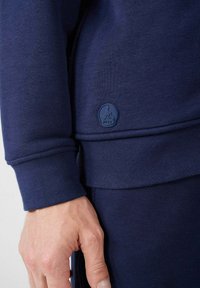 Sweatshirt bleu marine avec une texture lisse, des poignets côtelés et un petit logo en relief sur l'ourlet. Main reposant sur le tissu.