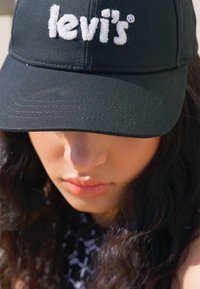 Melna kokvilnas cepure ar baltu izšūtu Levi's logo priekšpusē. Dizains ietver izliekamu malas daļu un strukturētu kroni.