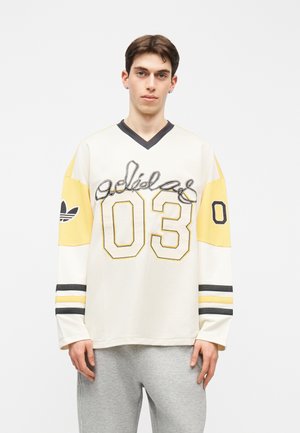 Joven de pie con una camiseta Adidas blanca y amarilla con "03" y el logo en las mangas, combinada con pantalones gris claro.