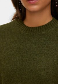 Femme portant un pull en maille vert foncé avec un col rond côtelé et de grandes boucles d'oreilles rondes foncées, gros plan sur le cou et les lèvres.