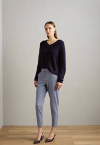 Marineblauwe textuurtrui, grijze aansluitende broek en zwarte pumps met spitse neuzen. Eenvoudige oorbellen aanvullen de outfit tegen een neutrale achtergrond.