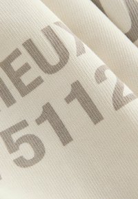 Tejido de algodón con fondo crema y grandes números y letras grises, que exhibe un diseño de superficie texturada y tejida.