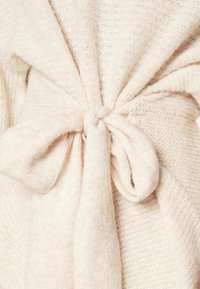 Écharpe tricotée beige avec une finition texturée. Dotée d'une ceinture nouée à la taille et d'un tissu doux et confortable dans une teinte claire et neutre.