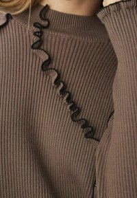 Pull côtelé marron avec un détail de volants ondulés noirs le long de l'encolure. La texture est douce et le tissu semble extensible.