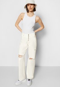 Top branco de alças, jeans brancos rasgados com cortes na frente, fecho de cinco botões, corte solto, combinado com ténis brancos e um chapéu de balde de tom claro.