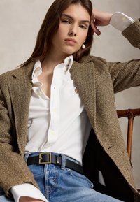 Joven con cabello castaño que lleva una camisa blanca abotonada, un blazer marrón de espiga, vaqueros azules y un pendiente de perlas, sentada con la mano apoyada en la cabeza.