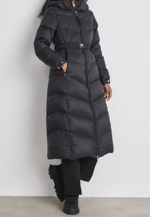 Długa czarna pikowana kurtka puchowa z paskiem w talii, zamkiem błyskawicznym i zatrzaskami na mankietach, noszona z czarnymi spodniami i masywnymi czarnymi butami.