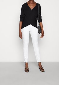 Schwarzer Wickelsweater mit langen Ärmeln, kombiniert mit weißen slim-fit Hosen und schwarzen Riemchen-Heels. Eine kleine schwarze Umhängetasche wird getragen.