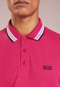 Rosa polo skjorta med en strukturerad tyg, som har en kontrasterande krage med svarta, vita och rosa ränder, samt en liten svart logotyp på bröstet.