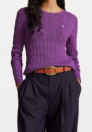 Femme portant un pull torsadé violet avec une ceinture marron et un pantalon taille haute foncé, mains dans les poches, sur fond blanc.