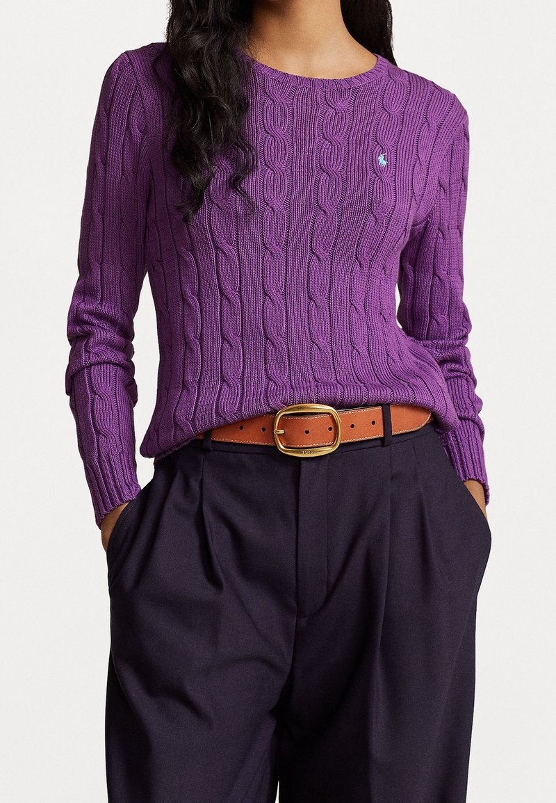 Femme portant un pull torsadé violet avec une ceinture marron et un pantalon taille haute foncé, mains dans les poches, sur fond blanc.