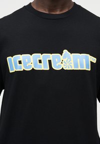 Μαύρο βαμβακερό μπλουζάκι με μεγάλο, πολυεπίπεδο γραφικό "παγωτού" σε μπλε και κίτρινο με σχεδιασμό στροβιλισμού. Στρογγυλή λαιμόκοψη και κοντά μανίκια.