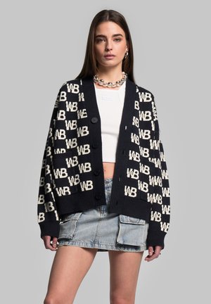 Cardigan blu navy con motivo crema "WB", dotato di chiusura a bottoni, polsini a coste e vestibilità ampia, indossato sopra una maglietta bianca e una gonna di jeans.