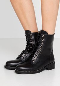 Černé kožené kotníkové boty s hladkým povrchem, kulatou špičkou, zlatými očky, šněrováním a nízkým blokovým podpatkem, s dekorativními hřebíky.