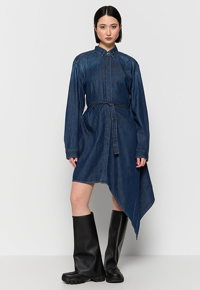 Robe en jean à manches longues, avec une fermeture à boutons sur le devant, une taille ceinturée et un ourlet asymétrique. Fabriquée en tissu bleu foncé avec une texture lisse.