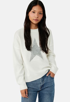 Ung kvinde med langt mørkt hår iført en hvid trøje med en stor grå stjerne og blå jeans, hånd i lommen, neutral ansigtsudtryk.