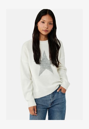 Ung kvinde med langt mørkt hår iført en hvid trøje med en stor grå stjerne og blå jeans, hånd i lommen, neutral ansigtsudtryk.