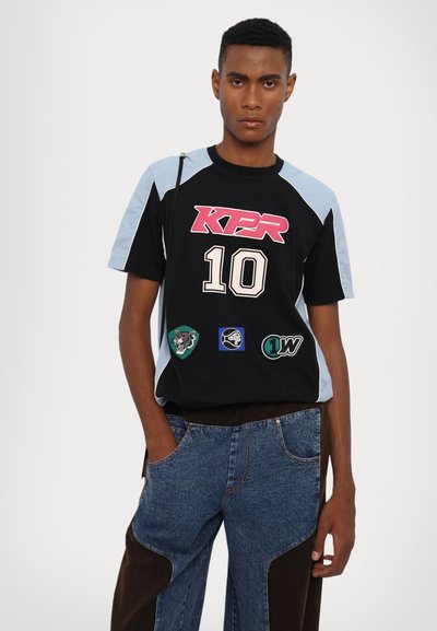 Camiseta de manga corta negra y azul claro con el logo "KPR", el número 10 y varios parches bordados. Combinada con pantalones de mezclilla azul.