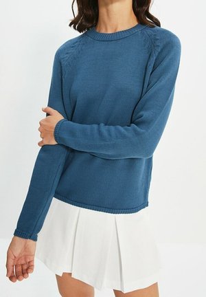 Vrouw met donker haar die een blauw, gebreide trui met lange mouwen en een witte geplooide rok draagt, staand met de ene arm over de andere gekruist.