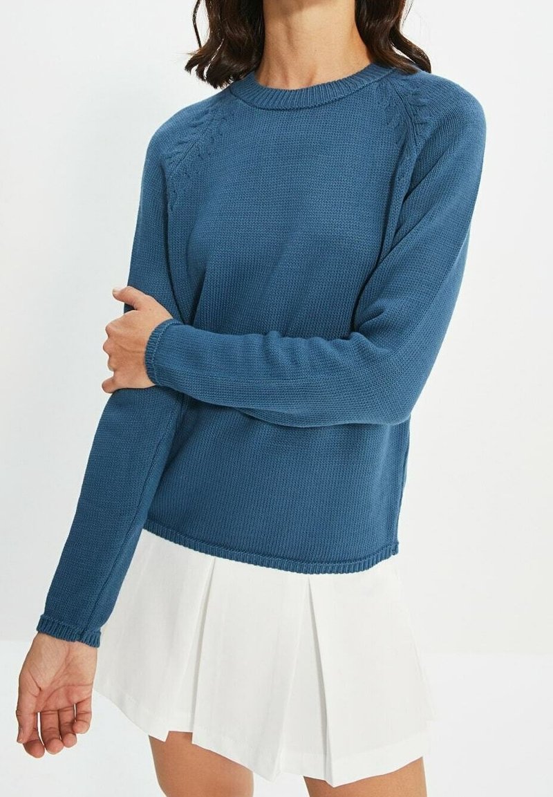 Femme aux cheveux foncés portant un pull bleu tricoté à manches longues et une jupe plissée blanche, debout avec un bras croisé sur l'autre.